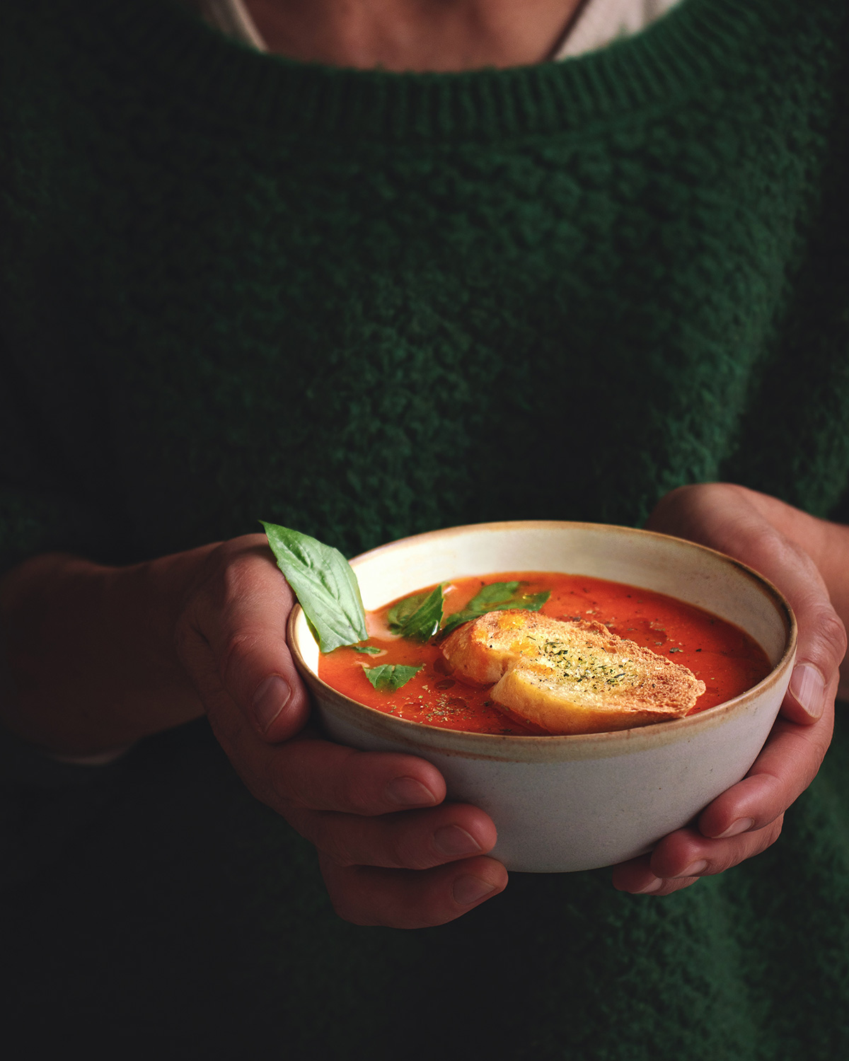 Fotografía de manos sosteniendo plato con sopa de tomate, pan tostado y albahaca